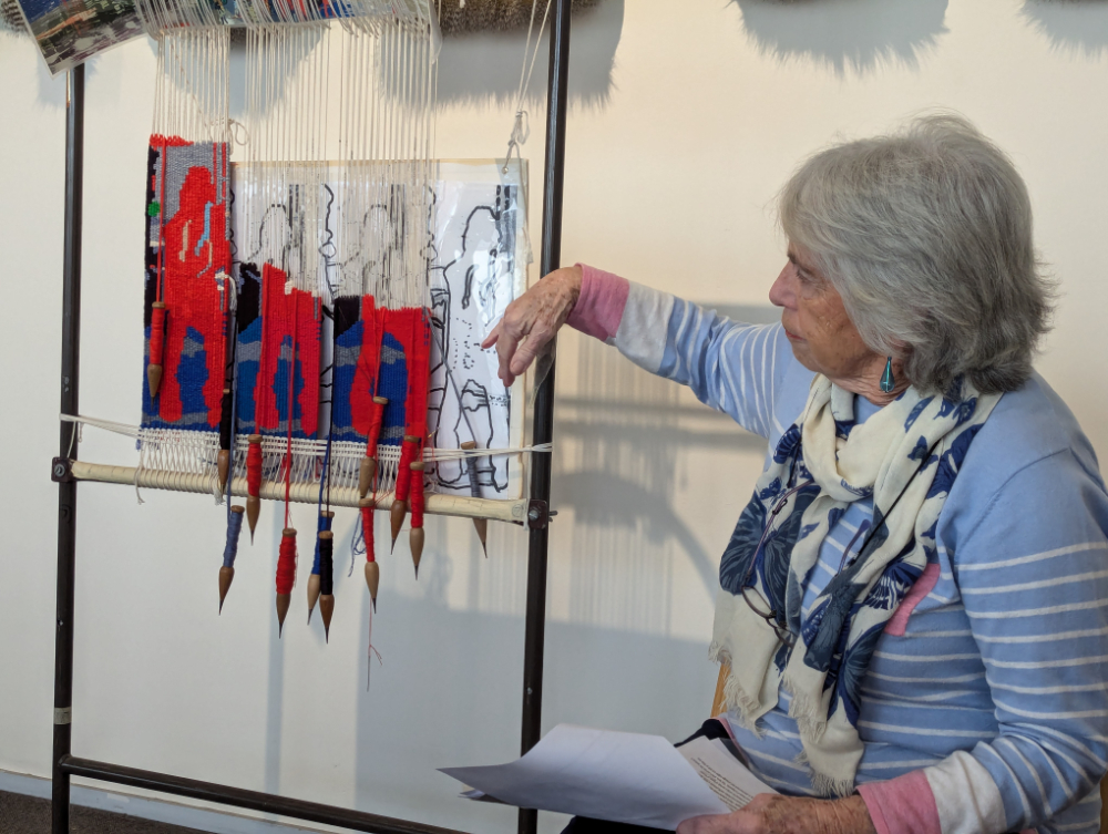 A woman presents a small tapestry art piece half finished on a loom